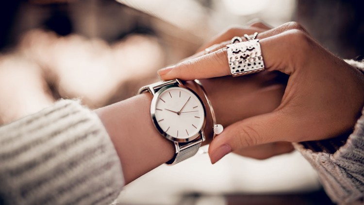 Eine silberne Armbanduhr mit weißem Zifferblatt wird an einem Handgelenk angepasst, im Hintergrund sind lichtdurchflutete Unschärfen.
