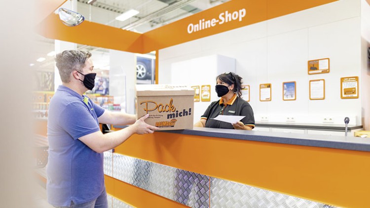Mann übergibt an einem Tresen im Globus Baumarkt Online-Shop eine große Kartonbox mit der Aufschrift „Pack mich!“.