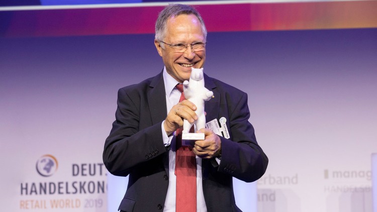 Person im Anzug mit roter Krawatte hält eine weiße Bärenfigur vor violettem Hintergrund mit Schriftzug „DEUTSCHER HANDELSKONGRESS“.