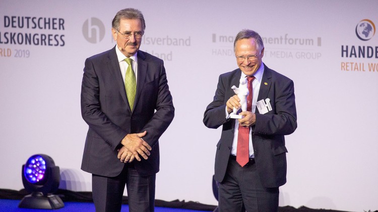 Zwei Männer in Anzügen auf einer Bühne, einer hält eine weiße Trophäe in den Händen, Schriftzug „DEUTSCHER HANDELSKONGRESS“ im Hintergrund.