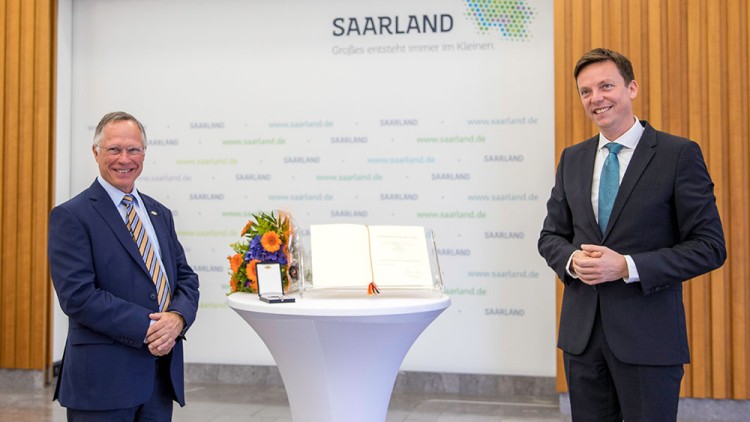 Stehtisch mit Blumenstrauß, Urkunde, Medaille und Hintergrundwand mit Schriftzügen „Saarland“ und Slogan „Großes entsteht im Kleinen“.