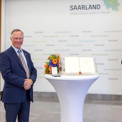 Stehtisch mit Blumenstrauß, Urkunde, Medaille und Hintergrundwand mit Schriftzügen „Saarland“ und Slogan „Großes entsteht im Kleinen“.