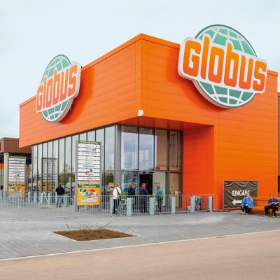 Eingangsbereich eines GLOBUS Marktes mit großem GLOBUS Logo, Glasfassade, Einkaufswagen und Personen auf dem Vorplatz.