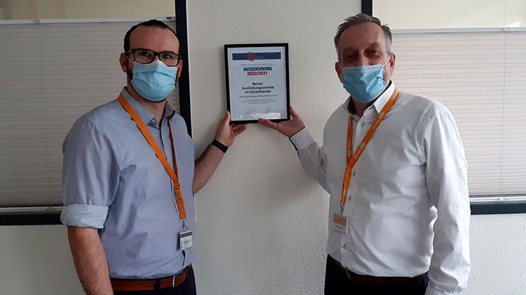 Zwei Personen in Hemden halten eine gerahmte Auszeichnung mit der Aufschrift „AUSZEICHNUNG 2020/2021“ vor einer Bürowand.