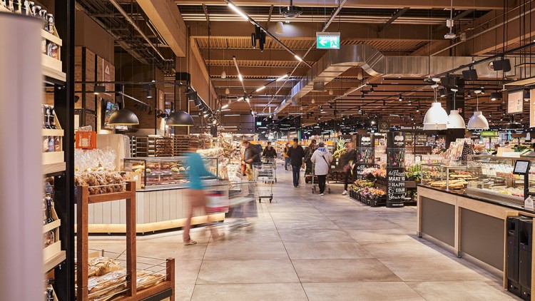 Ein GLOBUS Markt mit Obst- und Backwarenabteilung, Regalen, Einkaufenden und beleuchtetem Wegweiser an der Decke.