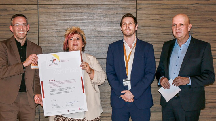 Vier Personen in Businesskleidung halten ein großes Zertifikat mit dem roten GLOBUS-Logo vor einer Holzvertäfelung.