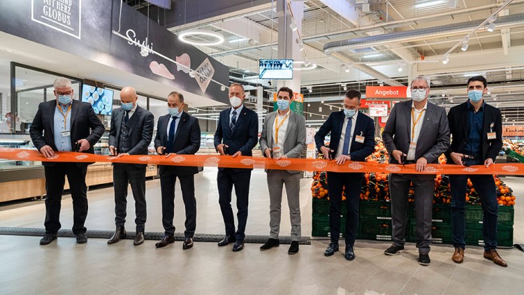 Mehrere Personen stehen in einem GLOBUS-Markt und schneiden ein oranges Band mit GLOBUS-Logo durch, im Hintergrund Obstkisten.