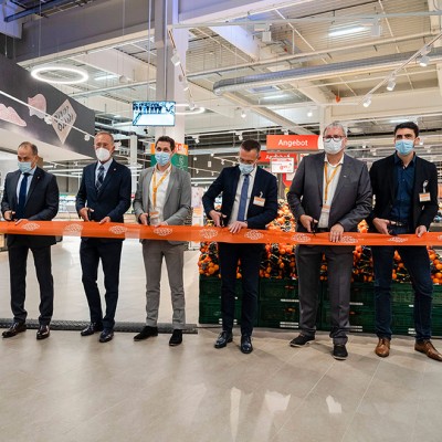Mehrere Personen stehen in einem GLOBUS-Markt und schneiden ein oranges Band mit GLOBUS-Logo durch, im Hintergrund Obstkisten.