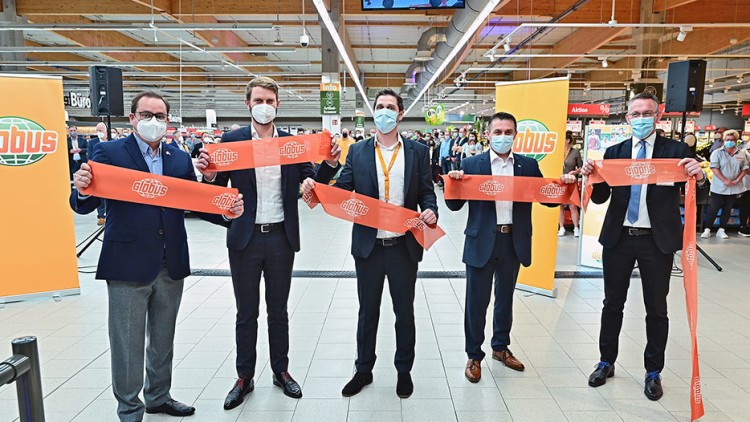 Fünf Personen in Anzügen halten ein rotes Band mit GLOBUS-Logo vor einem Publikum und GLOBUS-Werbetafeln.