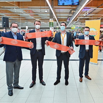 Fünf Personen in Anzügen halten ein rotes Band mit GLOBUS-Logo vor einem Publikum und GLOBUS-Werbetafeln.