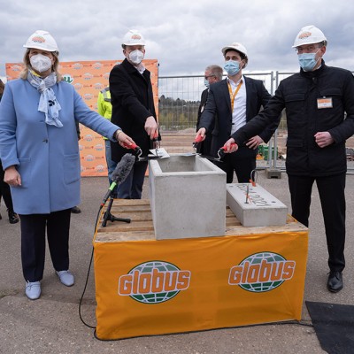 Personen in Schutzhelmen mit GLOBUS-Logo halten Kellen über Grundsteine bei einer Veranstaltung vor einer Baustelle.