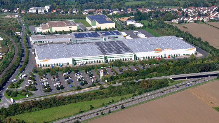 Luftaufnahme eines großen Gebäudekomplexes mit dem Schriftzug GLOBUS LOGISTIK, Parkplatz und umliegenden Feldern.