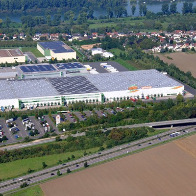 Luftaufnahme eines großen Gebäudekomplexes mit dem Schriftzug GLOBUS LOGISTIK, Parkplatz und umliegenden Feldern.