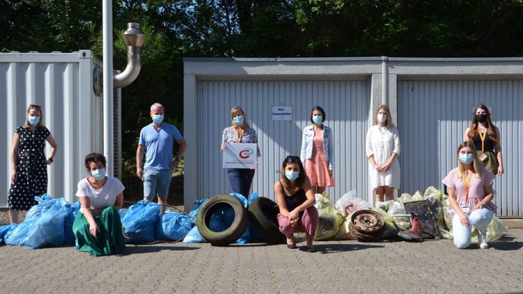 Personen stehen und sitzen vor einer Reihe von Müllsäcken, Reifen und Metallgegenständen, im Hintergrund sind Garagen zu sehen.