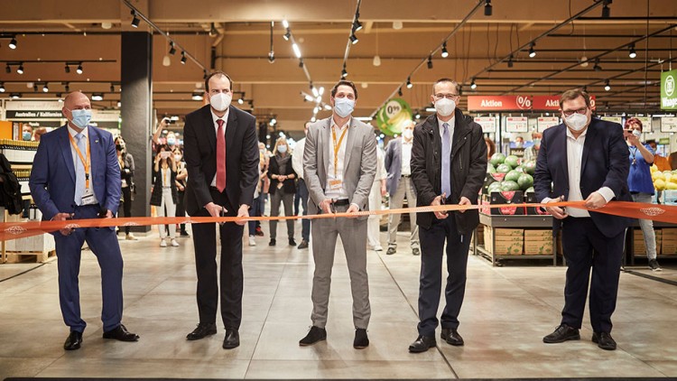 Fünf Personen schneiden ein orangenes Band in einem GLOBUS Markt durch, umgeben von Regalen mit Obst und Zuschauenden.