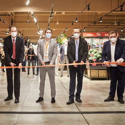 Fünf Personen schneiden ein orangenes Band in einem GLOBUS Markt durch, umgeben von Regalen mit Obst und Zuschauenden.