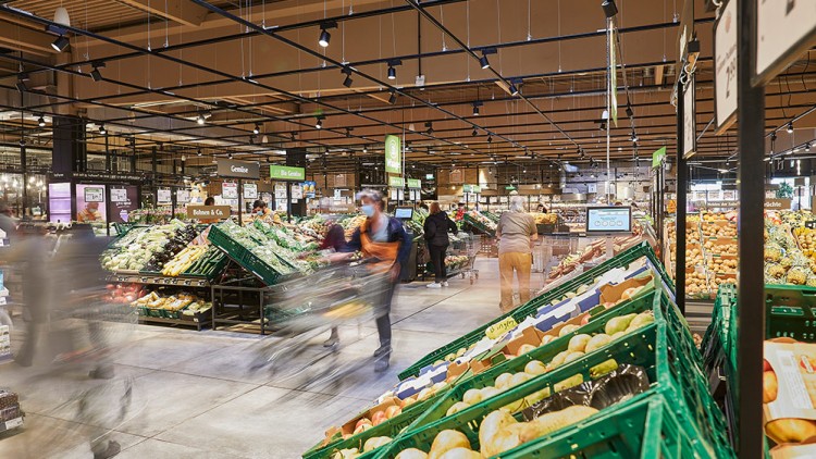 Gemüseabteilung in einem GLOBUS Markt mit grünen Kisten voller frischem Obst und Gemüse, Kunden mit Einkaufswägen im Gang.