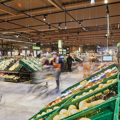 Gemüseabteilung in einem GLOBUS Markt mit grünen Kisten voller frischem Obst und Gemüse, Kunden mit Einkaufswägen im Gang.