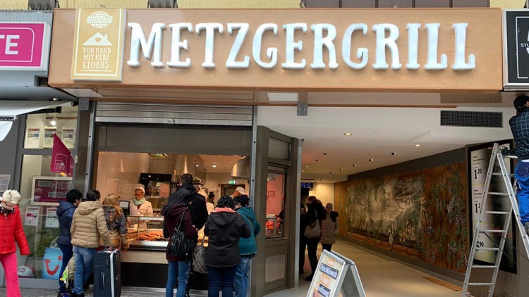 Mehrere Personen stehen vor einer Theke im METZGERGRILL von GLOBUS, über dem Eingang ein Schild mit „VON HAND MIT HERZ“.