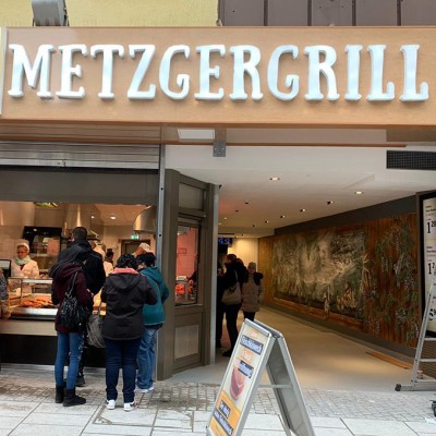 Mehrere Personen stehen vor einer Theke im METZGERGRILL von GLOBUS, über dem Eingang ein Schild mit „VON HAND MIT HERZ“.