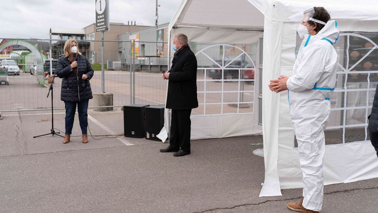 Eine Person spricht in ein Mikrofon, daneben stehen zwei weitere Personen vor einem Zelt auf einem Parkplatz.