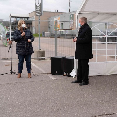 Eine Person spricht in ein Mikrofon, daneben stehen zwei weitere Personen vor einem Zelt auf einem Parkplatz.