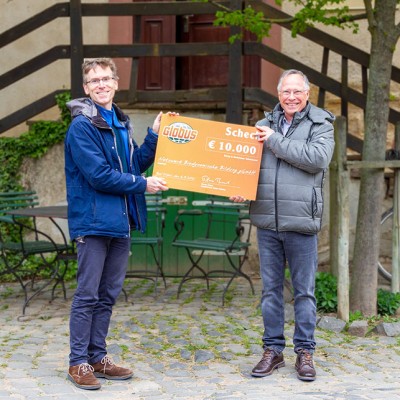 Zwei Personen halten einen großen Scheck mit GLOBUS-Logo und der Aufschrift „10.000 €“ vor einer Außenkulisse.