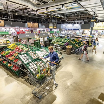 Eine Person mit Einkaufswagen steht vor Gemüseauslagen in einem GLOBUS Markt, über dem Bereich hängt ein Schild mit „Waage“.