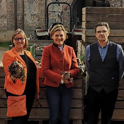 Fünf Personen stehen vor Holzkisten neben einem Traktor, zwei halten Wurzelgemüse mit Blättern in den Händen.