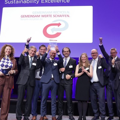 Eine Gruppe festlich gekleideter Personen feiert auf einer Bühne mit dem Schriftzug „Gemeinsam recyceln, gemeinsam Werte schaffen“.