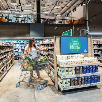 Eine Person schiebt einen Einkaufswagen mit frischen Lebensmitteln durch einen GLOBUS Markt, neben einem Regal mit der Aufschrift GUTfühl MARKT.