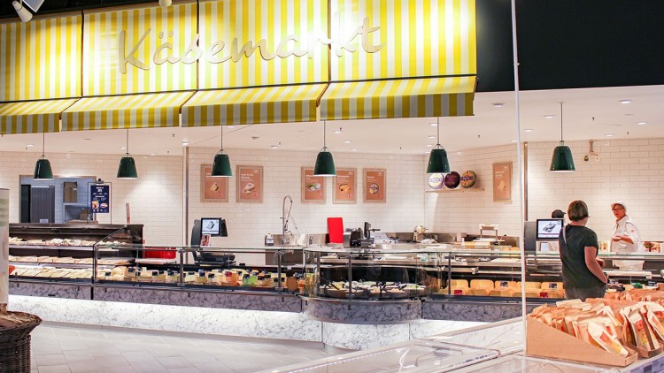 Bedientheke in einem Käsemarkt mit gelb-weiß gestreifter Markise, grünen Hängelampen und Käsewaren in Glasvitrinen.