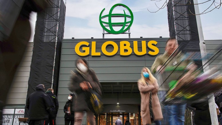 Menschen mit Einkaufswagen vor einem GLOBUS-Eingang mit großer gelber Schrift und grünem Logo auf grauer Fassade.