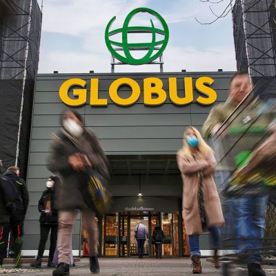 Menschen mit Einkaufswagen vor einem GLOBUS-Eingang mit großer gelber Schrift und grünem Logo auf grauer Fassade.