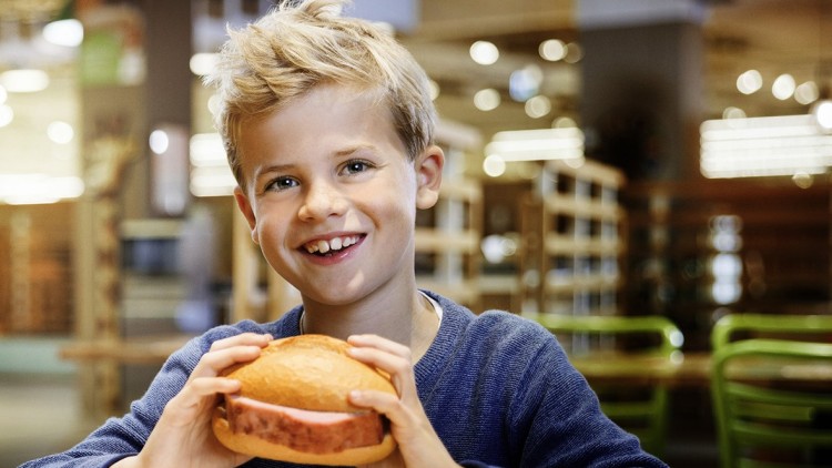 Person in blauer Kleidung hält ein belegtes Brötchen mit Wurst in den Händen, im Hintergrund unscharfes Ladeninterieur.