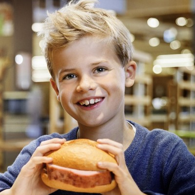 Person in blauer Kleidung hält ein belegtes Brötchen mit Wurst in den Händen, im Hintergrund unscharfes Ladeninterieur.