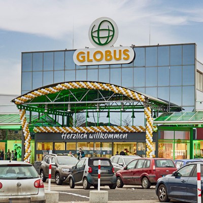 Eingang eines GLOBUS Marktes mit gold-weißer Ballongirlande und Schriftzug \"Herzlich willkommen\", Parkplatz mit Autos davor.