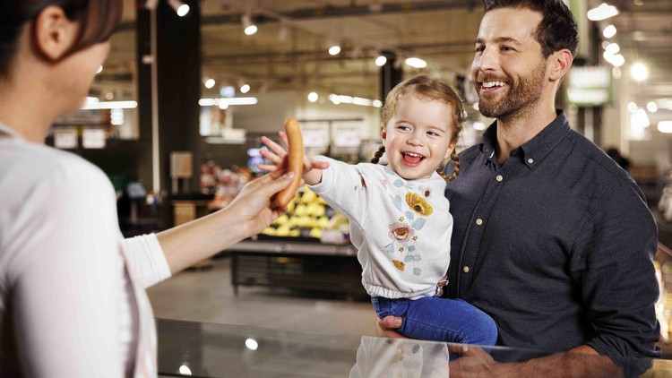 Ein Kind in gemusterter Kleidung nimmt eine Wurst aus der Hand einer Person, während es von einem Erwachsenen gehalten wird.