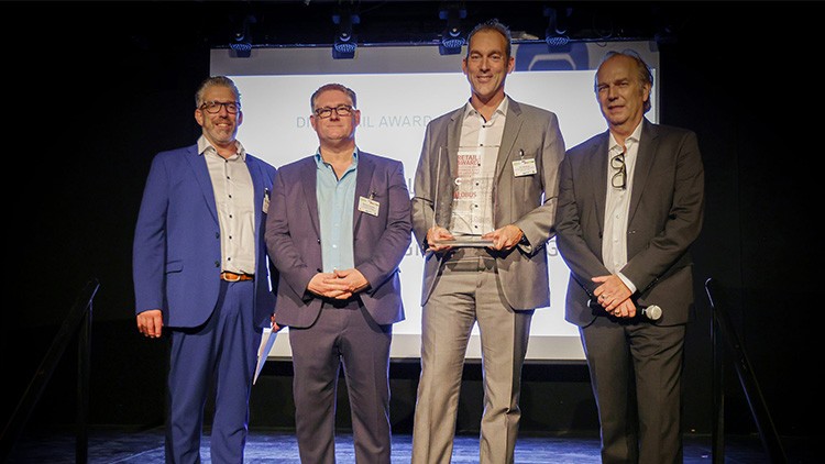Fünf Personen in Anzügen stehen auf einer Bühne vor einer Leinwand mit unleserlichem Text, einer hält eine Glas-Trophäe.
