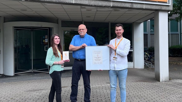 Drei Personen stehen vor einem Gebäude unter einem Banner mit der Aufschrift „Herzlich willkommen“ und halten eine Urkunde.