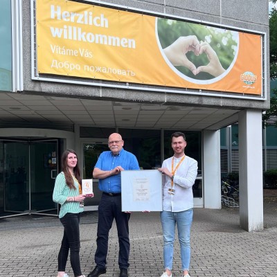 Drei Personen stehen vor einem Gebäude unter einem Banner mit der Aufschrift „Herzlich willkommen“ und halten eine Urkunde.