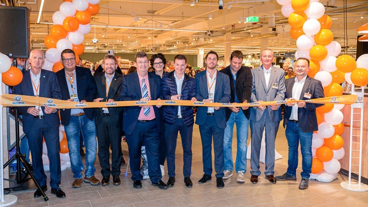 Mitarbeitende stehen unter einem orangenen und weißen Luftballonbogen und halten ein Band mit GLOBUS-Logo zur Eröffnung.