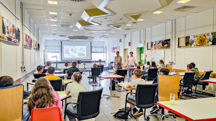 Ein heller Seminarraum mit Personen an Tischen, zwei Vortragende vor einer Projektion, an den Wänden hängen Poster.