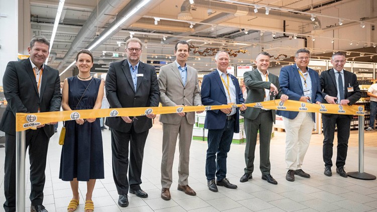 Mehrere Personen stehen hinter einem gelb-orangefarbenen Band mit dem GLOBUS-Logo in einer hell beleuchteten Verkaufshalle.