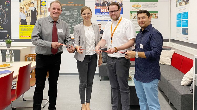 Vier Personen stehen zusammen in einem Bürobereich mit einer „Job Wall“ im Hintergrund und halten gemeinsam eine rote Schleife.