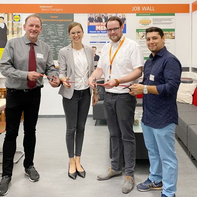Vier Personen stehen zusammen in einem Bürobereich mit einer „Job Wall“ im Hintergrund und halten gemeinsam eine rote Schleife.