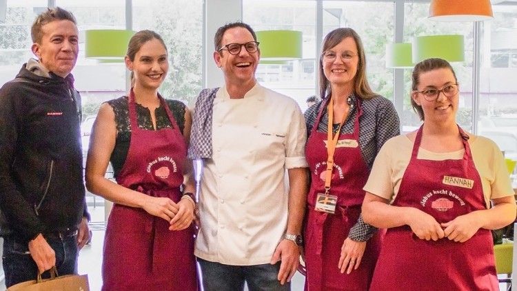 Mehrere Personen stehen in einer Küche, davon tragen vier bordeauxrote Schürzen mit dem Schriftzug „Globus kocht besser“.