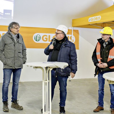 Drei Personen stehen vor einem GLOBUS-Banner, eine Person spricht mit Mikrofon, im Hintergrund ein gelbes GLOBUS-Zelt.
