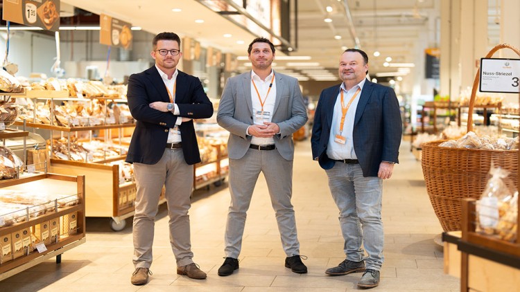Drei Personen in Businesskleidung stehen in einer gut beleuchteten Backwarenabteilung mit Brot und Gebäck auf Regalen.