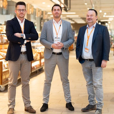Drei Personen in Businesskleidung stehen in einer gut beleuchteten Backwarenabteilung mit Brot und Gebäck auf Regalen.